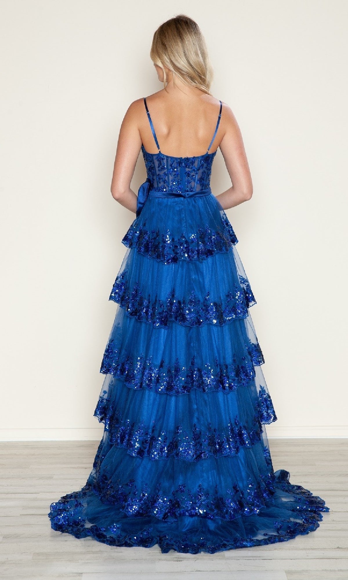 A woman stands with her back to the camera, wearing the Poly USA 9626 long prom dress—an A-line gown with a blue tiered skirt, shimmering sequin details, and delicate spaghetti straps—set against a plain, light-colored wall.